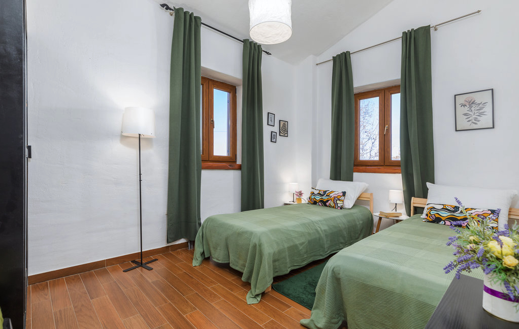 A cozy bedroom featuring two twin beds with green bedding, framed artwork on the white walls, and large windows adorned with green curtains. Soft lighting enhances the inviting atmosphere.